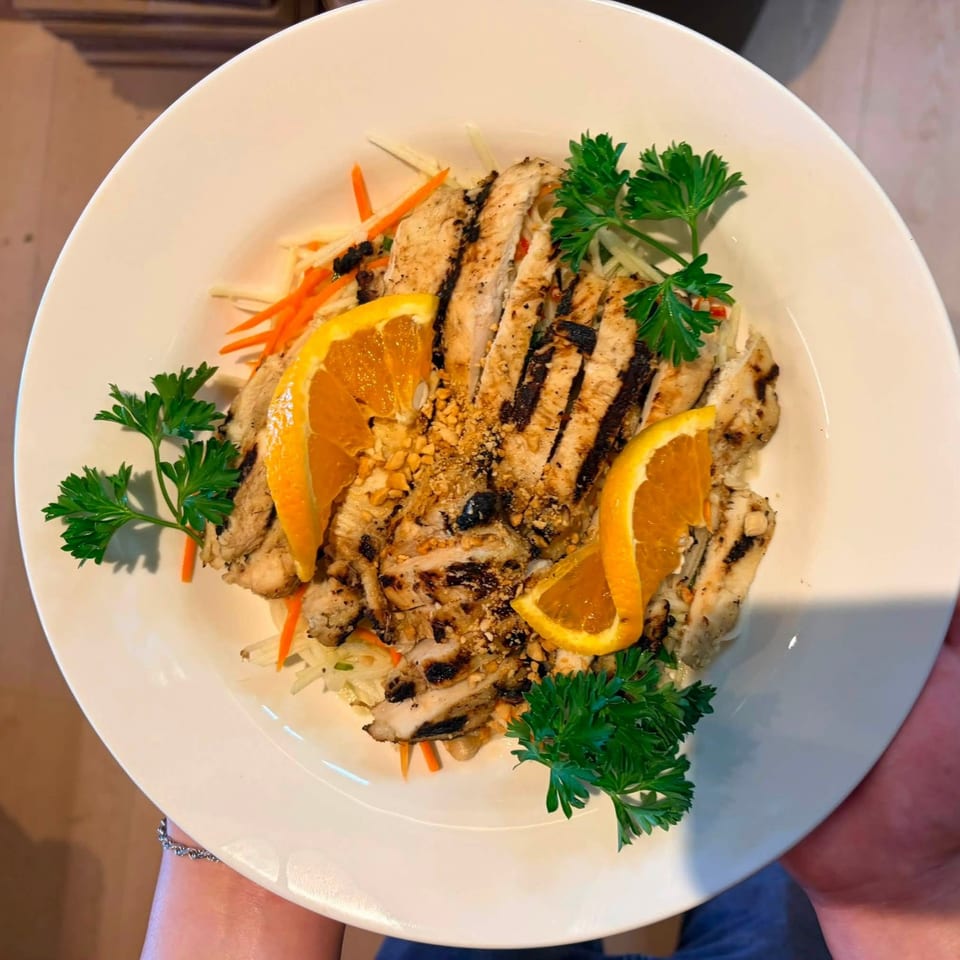 Grilled chicken salad with orange slices, peanuts, parsley, and shredded vegetables on a white plate.