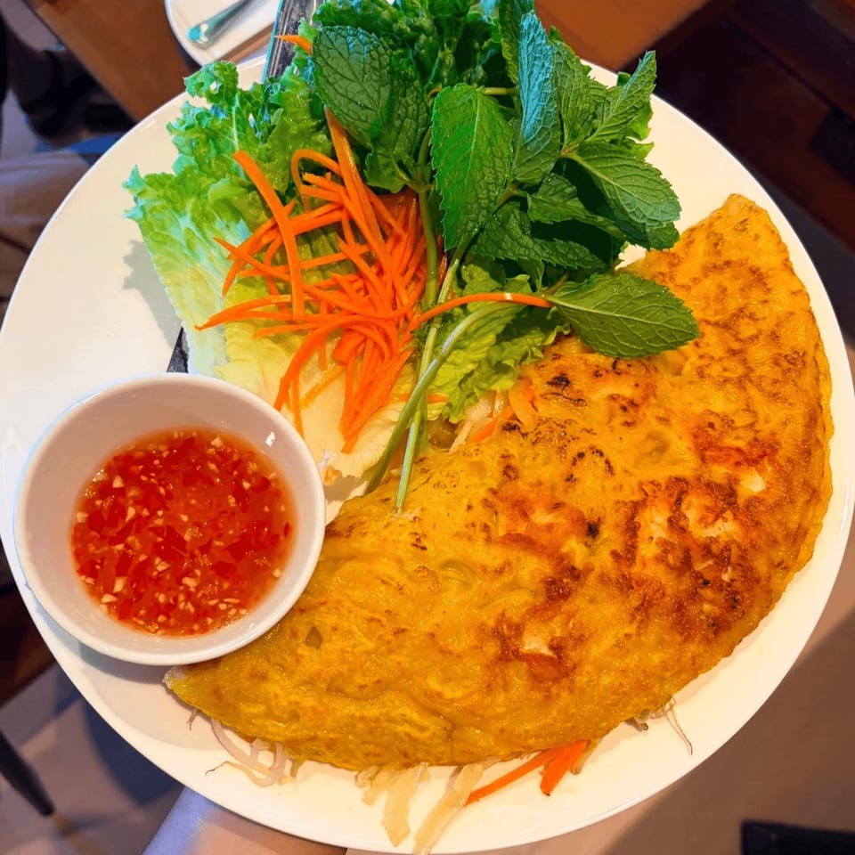 A golden Vietnamese savory crepe (Banh Xeo) with fresh herbs, shredded carrots, and chili dipping sauce.
