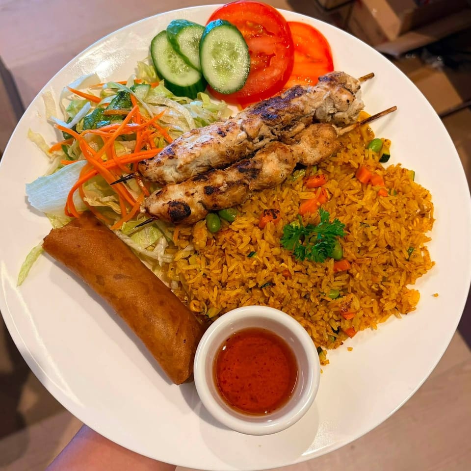 Chicken skewers with fried rice, a spring roll, salad, and dipping sauce on a white plate.